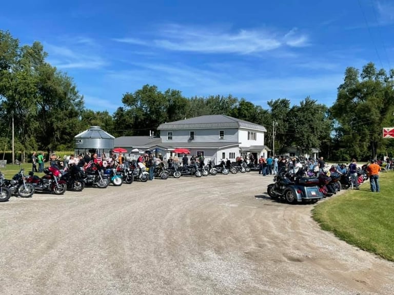 Large outdoor motorcycle rally at a community building with dozens of motorcycles parked on dirt grounds and crowds of people gathered on a sunny day