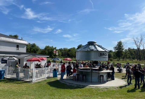 Outdoor event with converted grain silo bar structure, crowd of guests, white fence enclosure, and farm buildings on green lawn