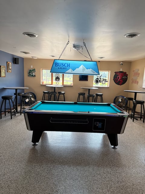 A black pool table with bright turquoise felt surface under a blue Busch Light neon sign in a recreational room with bar seating and windows