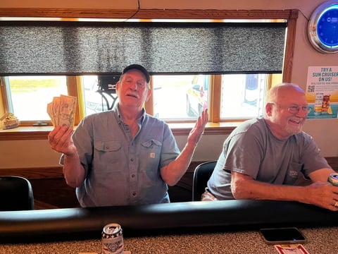 Two men sitting at a bar counter, one gesturing with raised hands wearing tan shirt, other man in gray shirt smiling, windows with blinds in background