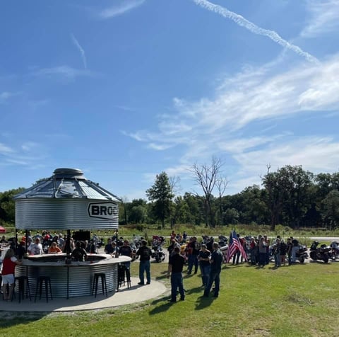 A large group of people gathered outdoors around a grain silo converted into a bar, with green grass and trees visible under a blue sky