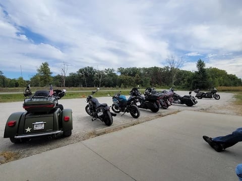 Group of motorcycles and a vintage hot rod car parked in a line on a rural paved area with green fields and trees in background