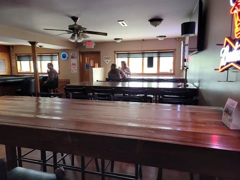 Interior of a dimly lit bar with wooden counter, ceiling fan, and staff behind the bar near windows