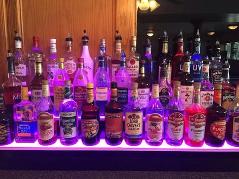 Bar display of spirits and liquor bottles illuminated with purple and magenta neon lighting against wooden wall panels
