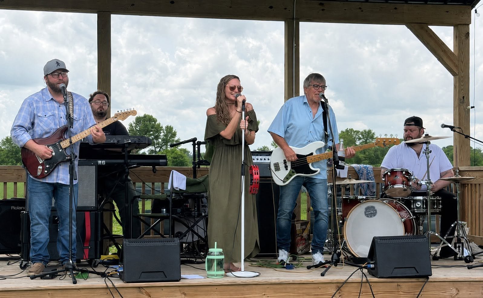 Live band performing on outdoor stage with guitarist, keyboardist, vocalist, bassist, and drummer under a covered pavilion