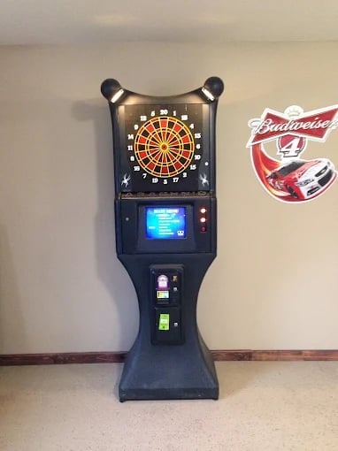 Electronic dartboard arcade game machine with lit display and controls, next to a Budweiser sign on a beige wall